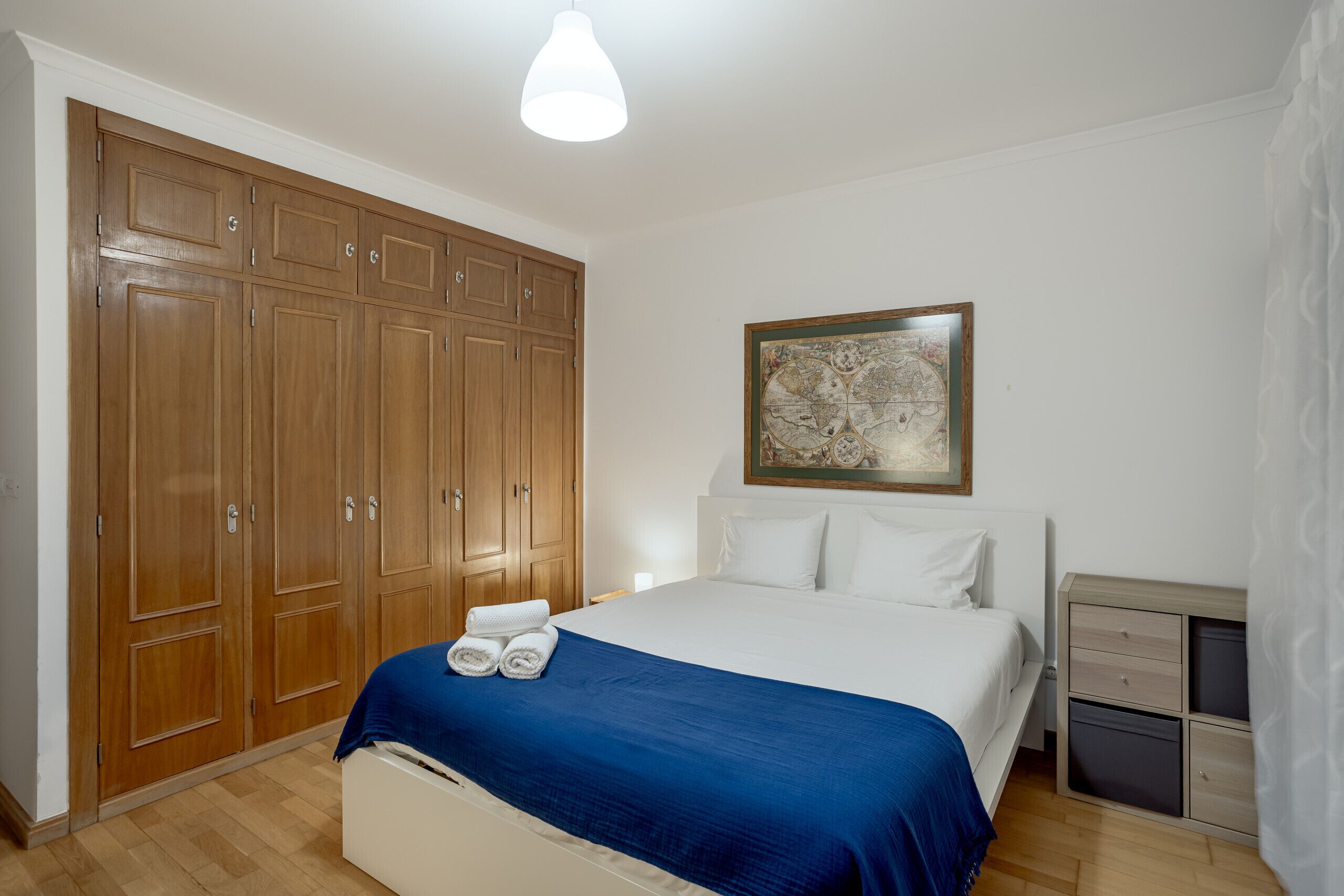 Ein Schlafzimmer mit großem Kleiderschrank, einem Bett mit blauer Decke, einem Nachttisch und einem gerahmten Kunstwerk an der Wand.