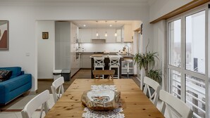 A modern kitchen with white cabinets, a dining table with a wooden centerpiece, and a view of the city through large windows. - Carcavelos Modern Apartment by Homing (Cascais)