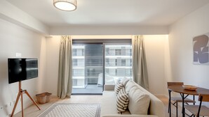A modern living room with a flat-screen TV, a sofa, a dining table, and a view through a large window. - Modern Lisbon W/ Balcony Pool II (Lisbon)