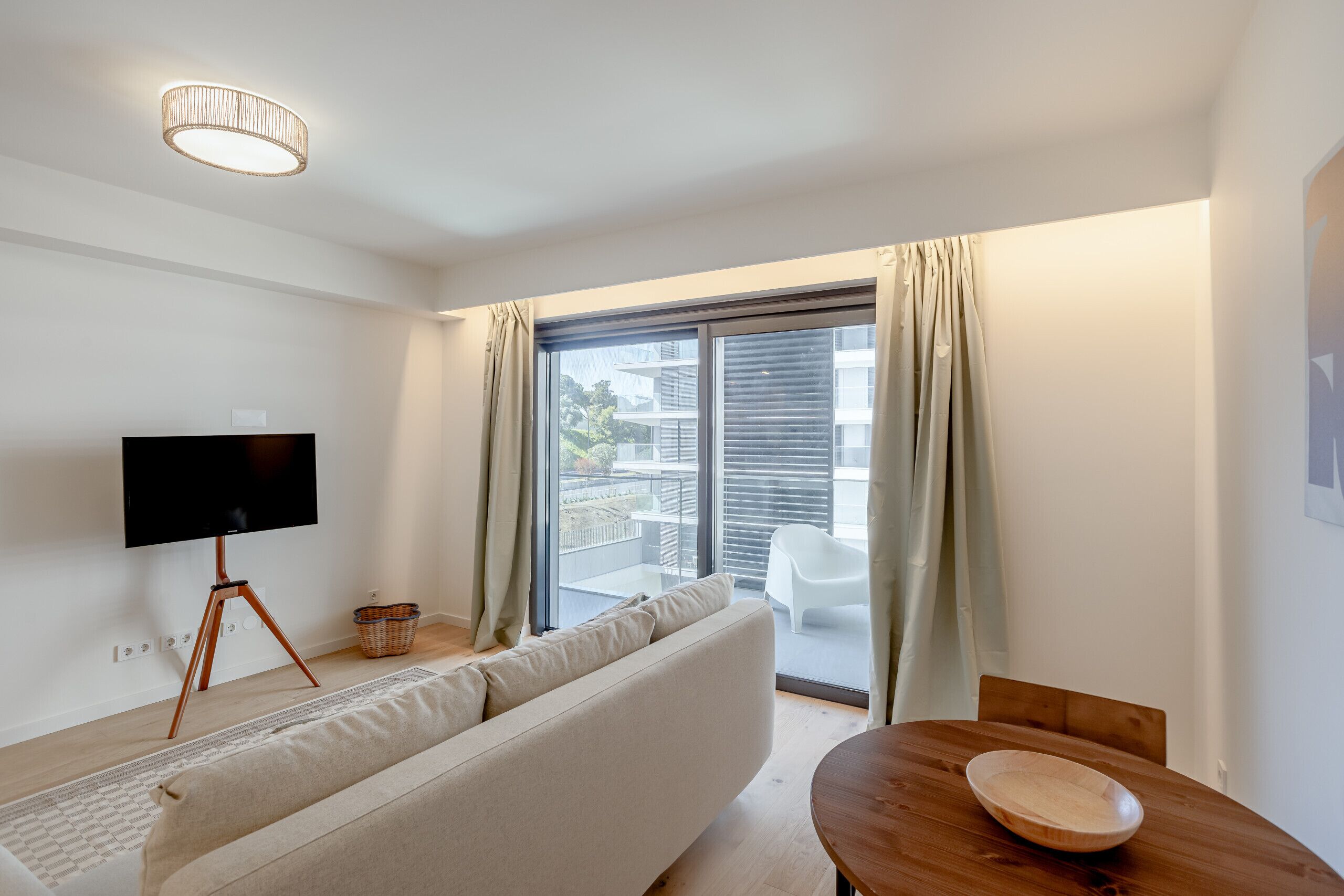 A modern living room with a sofa, a wooden table, a flat-screen TV on a tripod stand, and a balcony with a view of buildings.