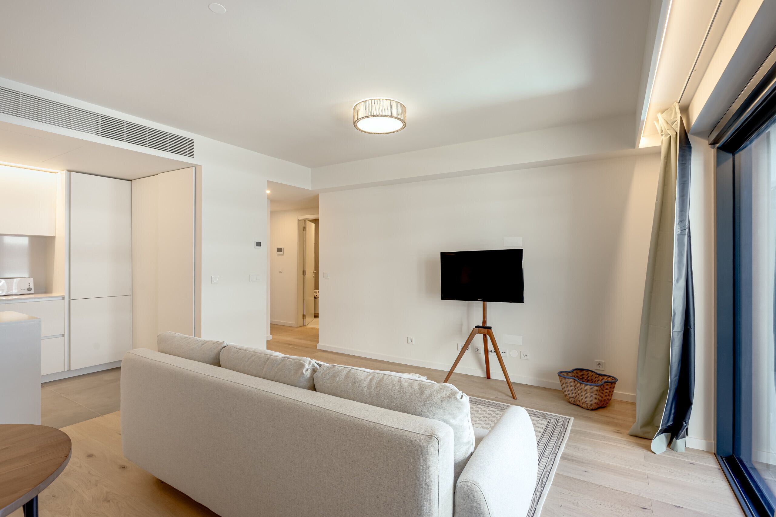 A modern living room with a sofa, a flat-screen TV on a tripod stand, a kitchen area with cabinets, and a large window with curtains.