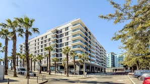 A multi-story hotel with a modern design, featuring balconies, a parking area, and palm trees. - Modern Lisbon W/ Balcony Pool II (Lisbon)