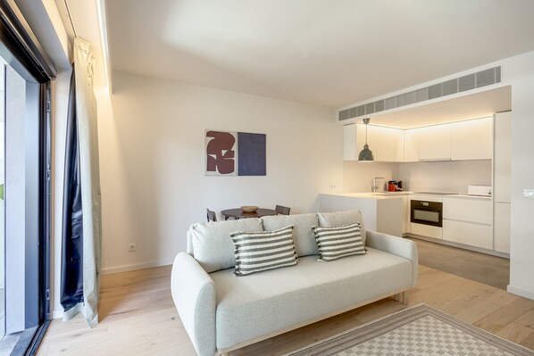 A modern living room with a sofa, a dining table, and a kitchen area with white cabinets and appliances. - Modern Lisbon W/ Balcony Pool II (Lisbon)