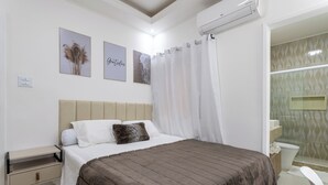 Un dormitorio con cama, mesita de noche, aire acondicionado instalado en la pared y un baño visible a través de una puerta abierta.