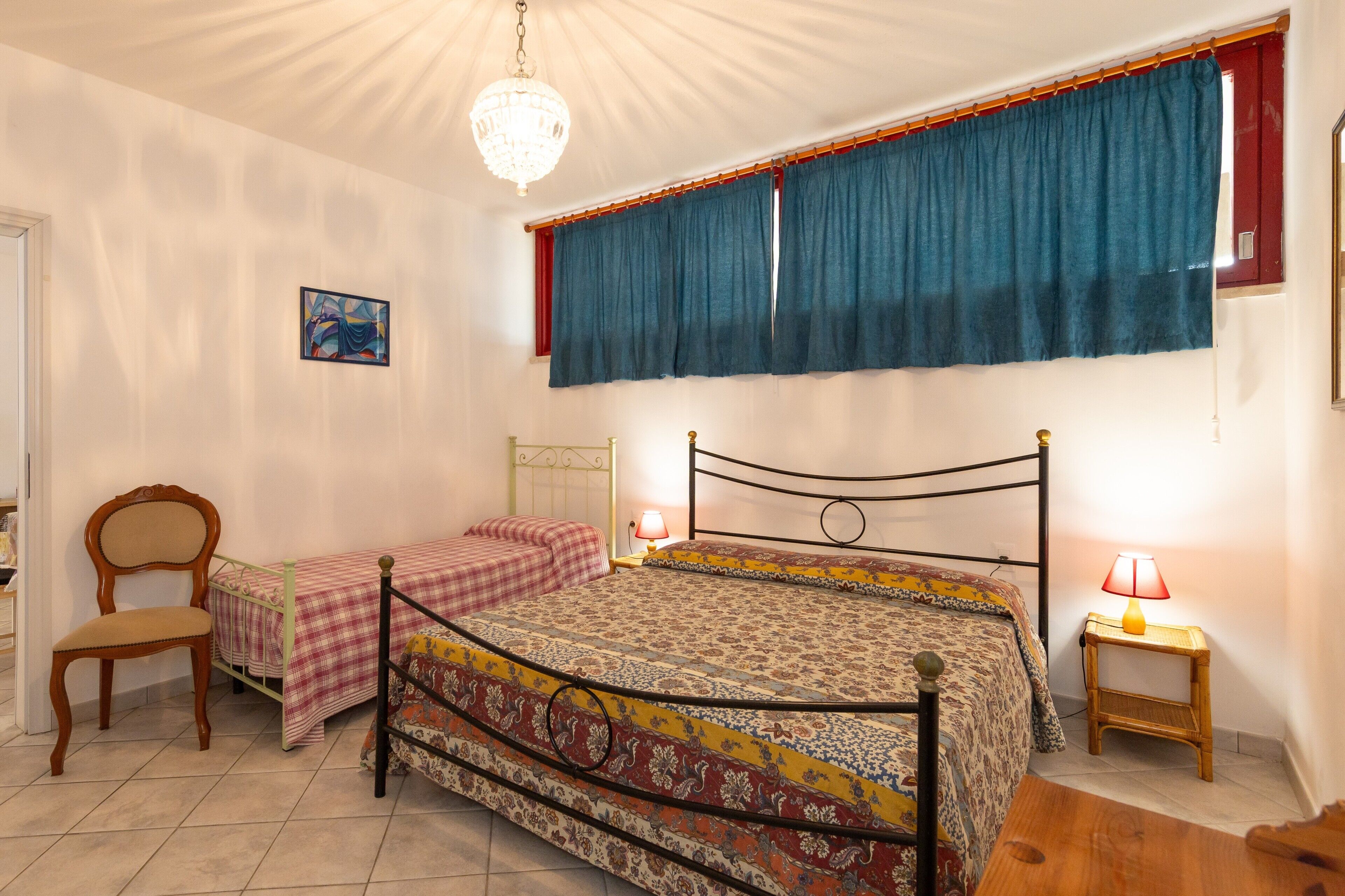 Une chambre à coucher comprenant un lit, une chaise, une table de chevet, une lampe et une fenêtre avec des rideaux.