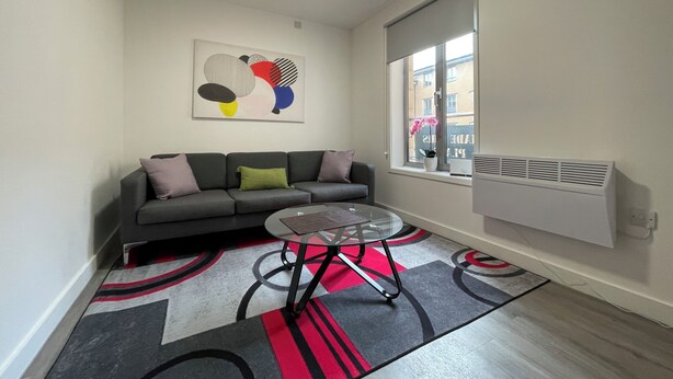 A modern living room with a grey sofa, a glass coffee table, and a large window with a view of buildings. - Downtown Sheffield Retreat (Sheffield)