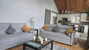 Un salon avec deux canapés gris, une table basse ornée de bougies et, au fond, une cuisine.