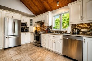 A modern kitchen with white cabinetry, stainless steel appliances, a stone backsplash, and a wooden ceiling. - Spa & Private beach by the Lake | Repère du Cerf (Saint-Côme)