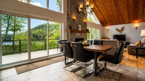 Une pièce spacieuse comprenant un coin repas, une cheminée, un téléviseur et un balcon offrant une vue sur les arbres et un lac.