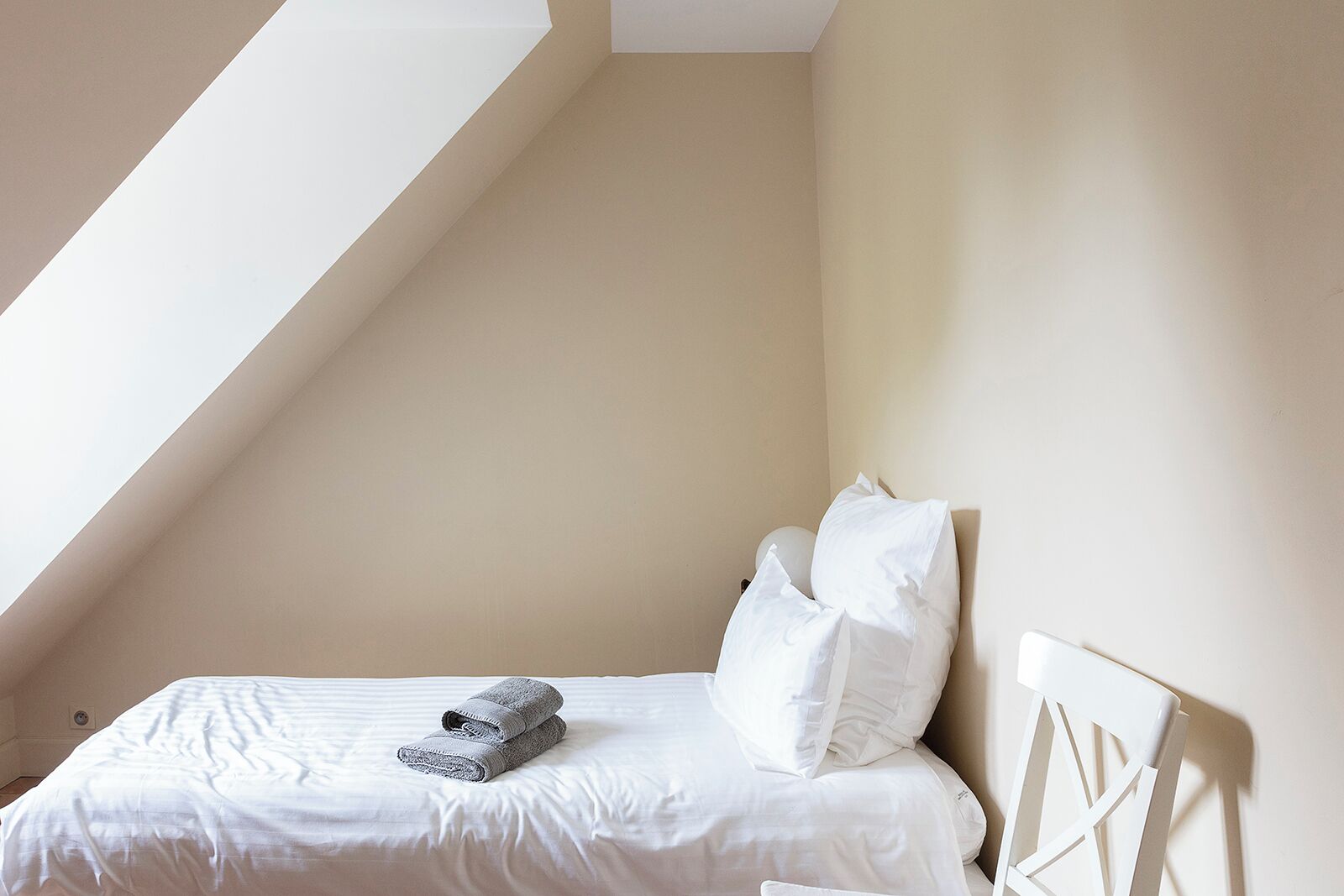 A bedroom with a sloped ceiling, a bed with white bedding and pillows, and a folded towel on the bed.