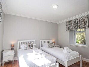 A bedroom with two beds, a window with patterned valance, a bedside table with a lamp, and a nightstand. - Craig Cottage (Banff)