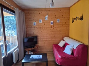 Una habitación acogedora con paredes de madera, un sofá rojo, un televisor sobre un mueble bajo y una ventana con cortinas.