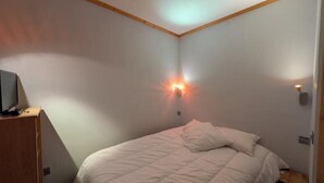 Una stanza piccola e arredata in modo essenziale, con un letto, una televisione su un supporto in legno e un punto luce a parete.