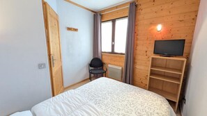 Un dormitorio con cama, una silla, un televisor sobre una estantería de madera y una ventana con cortinas.
