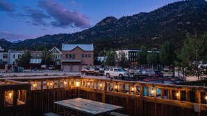 A wooden deck with tables and chairs, a parking lot with cars, and buildings in the background. - Riverwalk Path! Heart of South Main w/ Peak Views (Buena Vista)