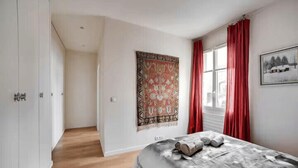 Une chambre à coucher avec un lit, une fenêtre ornée de rideaux rouges, une tapisserie accrochée au mur et un couloir.