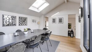 Un espace de salle à manger moderne, avec une longue table, des chaises en osier et des œuvres d’art encadrées accrochées aux murs.