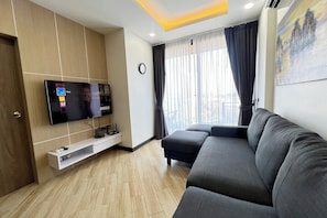 Ein modernes Wohnzimmer mit einem flachbildfernseher an der Wand, einem dunkelgrauen Sofa, einem Fenster mit transparenten Vorhängen und einem Gemälde an der Wand.