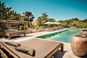 Un área junto a la piscina con sillones de descanso, una pérgola de madera y una piscina de color azul cristalino.