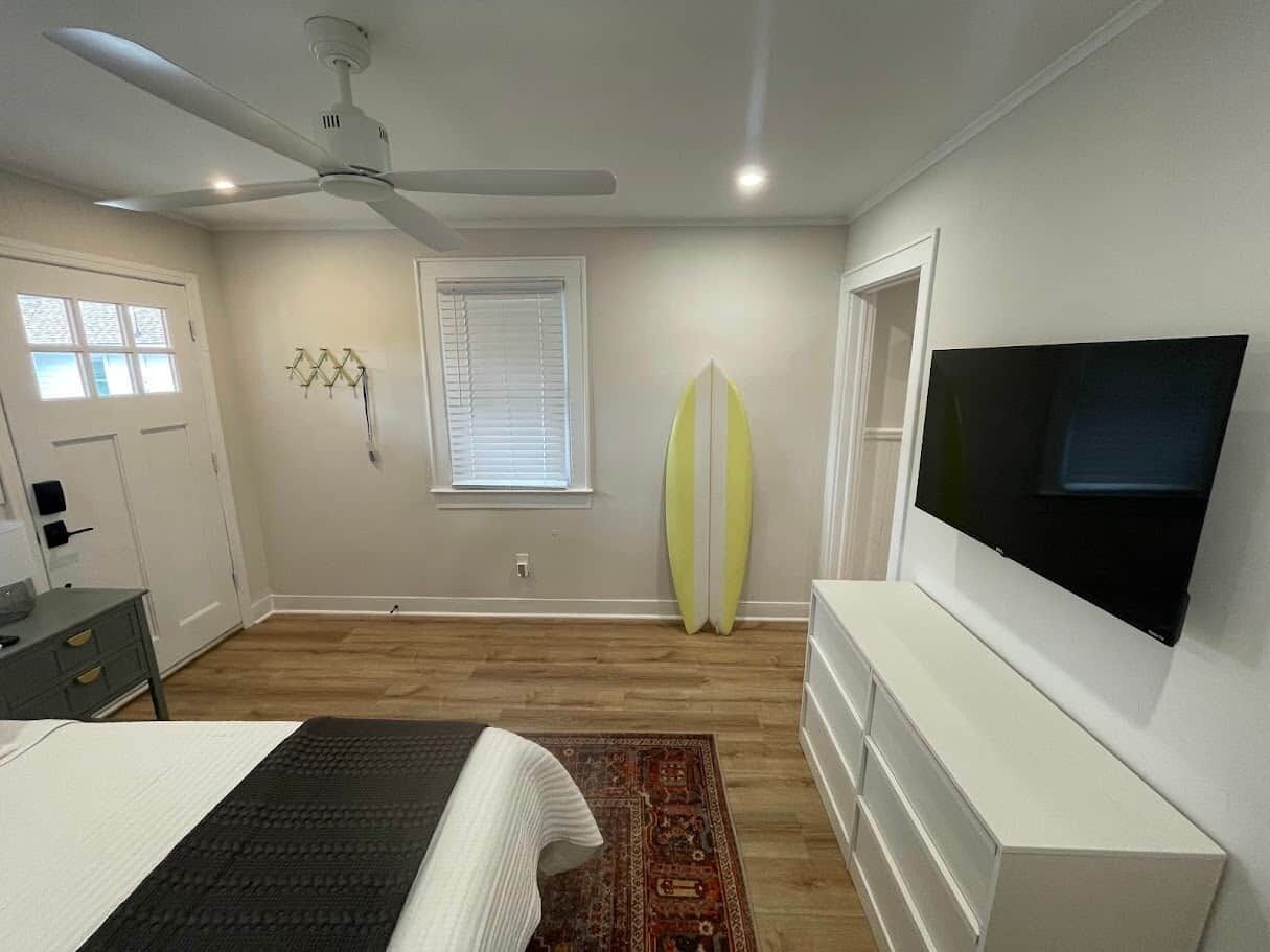 A bedroom with a bed, a ceiling fan, a TV, a surfboard, and a window.