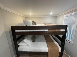 Una cama litera con almohadas y una colcha, una ventana con persianas y una lámpara en el techo.