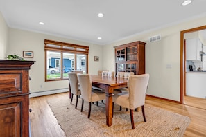 Une salle à manger avec une table en bois, six chaises, un tapis, une fenêtre avec des stores et une cuisine visible à travers une porte ouverte.