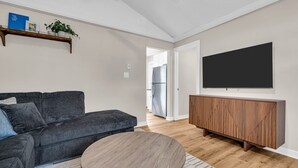 Een woonkamer met een donkergrijze bank, een ronde houten salontafel, een houten kast en een grote flatscreen televisie aan de muur.