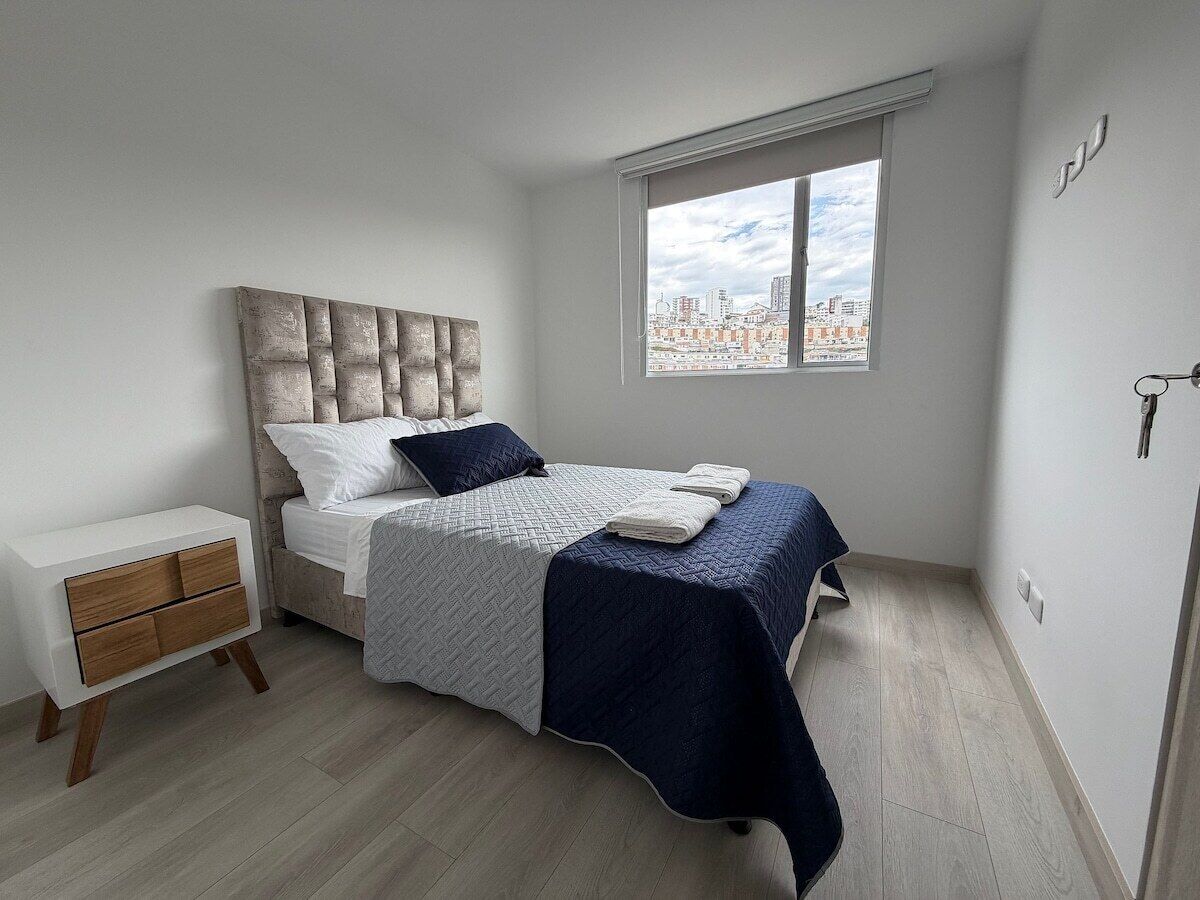 Un dormitorio con cama, mesita de noche, ventana y vista a la ciudad, además de una puerta.