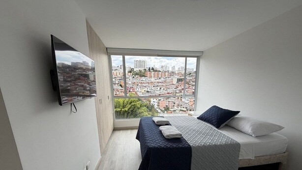 Un dormitorio con cama, televisor montado en la pared y un amplio ventanal con vistas a la ciudad.