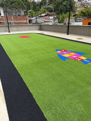 Uma área com grama sintética verde, um jogo de amarelinha colorido e uma pista de corrida preta.