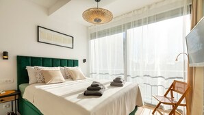 Ein Hotelzimmer mit Bett, Sessel, Fernseher und einem Fenster mit durchsichtigen Vorhängen.