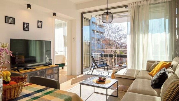 Ein modernes Wohnzimmer mit Flachbildfernseher, einer Couch, einem Couchtisch und einem Balkon mit Blick auf Gebäude.
