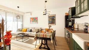 Ein modernes Wohnzimmer mit Sofa, Couchtisch und Esstisch. Es gibt eine Küchenzeile mit Schränken und einer Spüle.