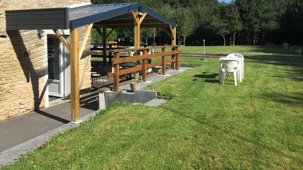 A covered outdoor seating area with tables and chairs, a stone building, and a grassy lawn. - Gite Eltiwen esprit bar (la croix hellean)