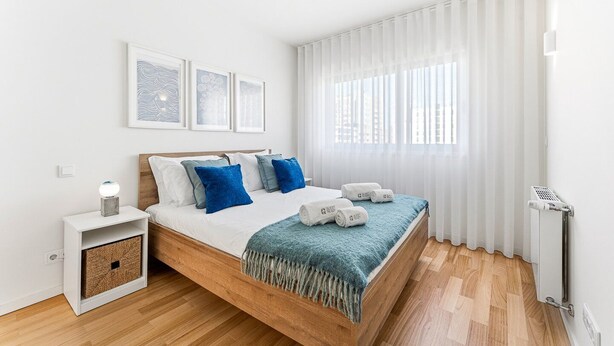 Un dormitorio moderno con una cama de madera, paredes blancas y un ventanal con cortinas traslúcidas.