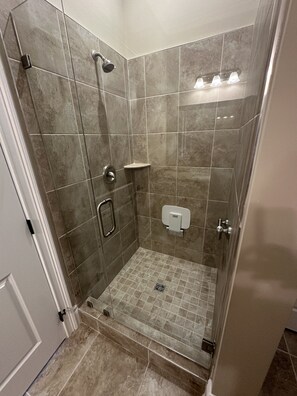 Una ducha a nivel con puerta de vidrio, paredes con azulejos, un cabezal de ducha manual, un control para la ducha manual, un dispensador de toallas y un desagüe en el suelo.