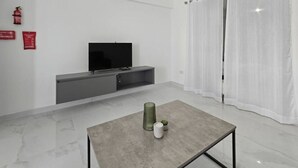 Ein modernes Wohnzimmer mit Flachbildfernseher, einer grauen Unterhaltungsecke, einem Glastisch und weißen Vorhängen.