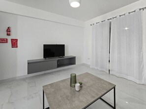 A modern living room with a flat-screen TV, a grey entertainment unit, a glass coffee table, and white curtains. - Centrall located modern 1 bedroom apartment with balcony (San Pawl il-Baħar)