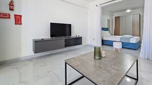 Ein modernes Hotelzimmer mit Flachbildfernseher, einer grauen Ablage, einem kleinen Tisch mit einer Vase, einem Bett mit blauer Bettwäsche und einer Schiebetür zu einem weiteren Raum.
