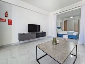 A modern hotel room with a flat-screen TV, a grey console, a small table with a vase, a bed with blue bedding, and a sliding door leading to another room. - Centrall located modern 1 bedroom apartment with balcony (San Pawl il-Baħar)