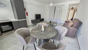 Ein modernes Wohnzimmer mit einem runden Tisch, grauen Sesseln, einer Couch, einem Fernseher und einer Küchenzeile mit Herd und Schränken.