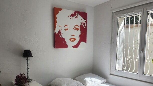A bedroom with a bed, a nightstand, a lamp, a ceiling fan, and a window with bars. - Cosy apartment (Nîmes)