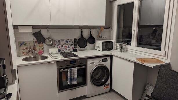 Ett kompakt kök med tvättmaskin, mikrovågsugn och ett fönster med persienner.