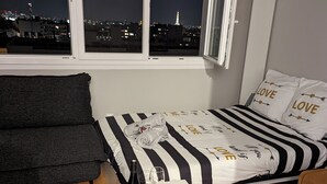 Ett välordnat sovrum med en säng som har ett randigt överkast, ett sängbord med en flaska och ett glas, och en utsikt över en stadssiluett genom ett fönster.