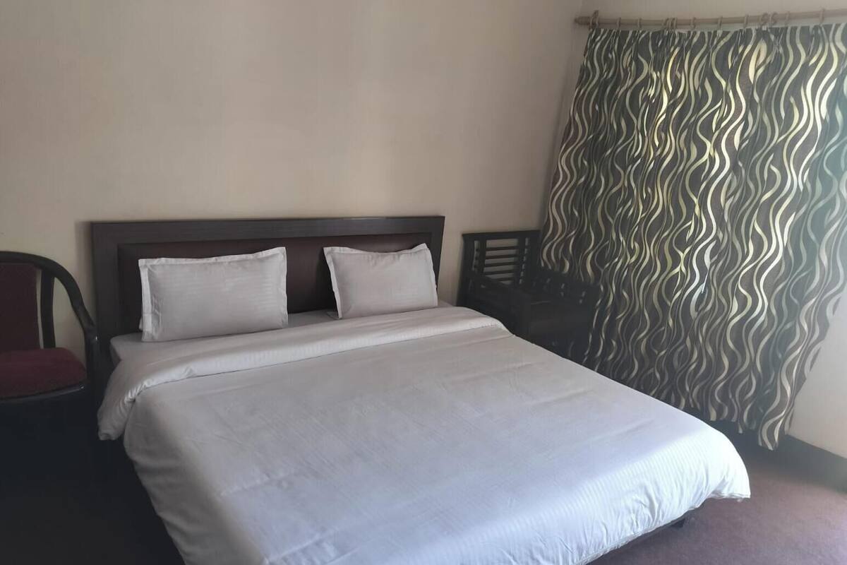 A hotel room with a bed, two pillows, a chair, and a curtain with a wavy pattern.
