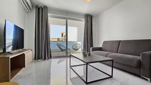 Ein modernes Wohnzimmer mit einer Couch, einem Couchtisch, einem Fernseher und einem Balkon mit Vorhängen.