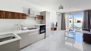 Eine moderne Küche mit Einbauofen, Kaffeemaschine und einem kleinen Tisch. Es gibt einen Essbereich mit Glastisch und einer Couch. Der Raum verfügt über ein großes Fenster mit Vorhängen.