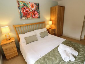 A neatly made bed with white linens, a green patterned bedspread, and two pillows. There is a wooden headboard, a nightstand with a lamp, and a wardrobe. A painting of flowers hangs above the bed. - 3 Rhodewood House (Saundersfoot)