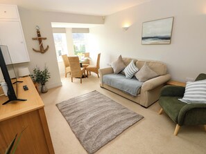 A living room with a beige sofa, a green armchair, a wooden desk with a computer, a dining area with wicker chairs, and a view through a large window. - 3 Rhodewood House (Saundersfoot)