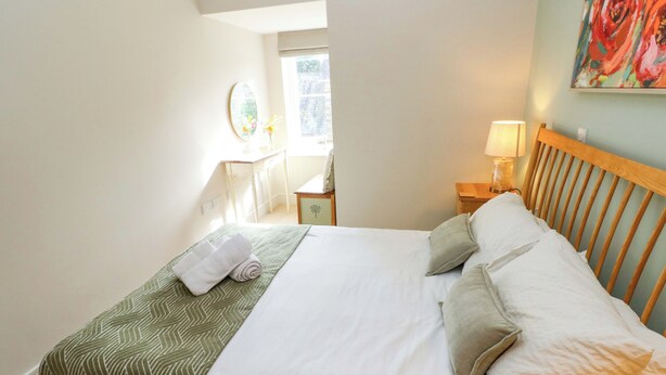 A neatly made bed with a green patterned bedspread, a white comforter, and two pillows. There is a wooden headboard, a bedside table with a lamp, and a wall-mounted painting. - 3 Rhodewood House (Saundersfoot)